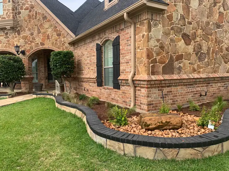 Curved stone bed edging with a boulder and river rock in front of a stone and brick home