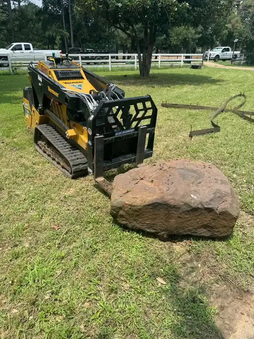 Yellow tracked skid steer parked beside a boulder on a residential lawn