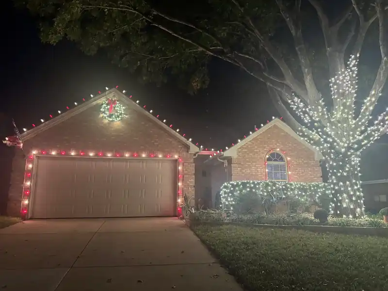 Night Christmas-light install on a brick home: red and white roofline lights and a white-lit oak tree