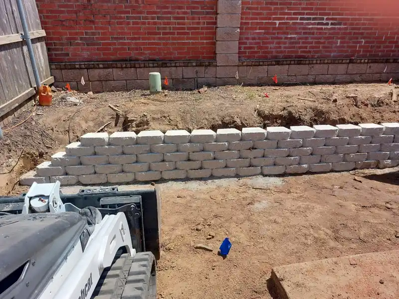 A retaining wall block course going in on a regraded slope, with a skid steer at the edge of the frame