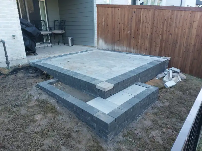 Paver patio with a charcoal border and two steps down to the lawn, built against a stained wood fence
